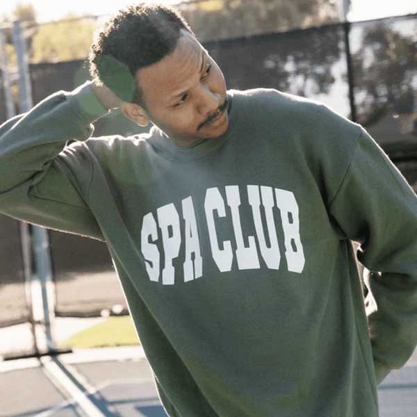Man outdoors wearing a green Spa Club Crew Neck Sweatshirt | Lucky Owl with white bold lettering, casually adjusting his hair on a sunny day near a fenced area.