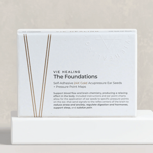 The Foundation - 24K Gold Ear Seed | Vie Healing box displayed on a white pedestal, featuring self-adhesive gold acupressure ear seeds with pressure point maps for wellness and stress relief.