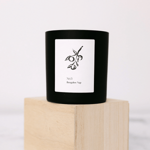 Black candle in a minimalist glass holder with a white label featuring an olive branch illustration, standing on a light wooden block, No. 3 Bungalow Nap (Fiji) Candle | Lucky Owl.