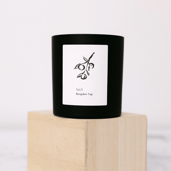 Black candle in a minimalist glass holder with a white label featuring an olive branch illustration, standing on a light wooden block, No. 3 Bungalow Nap (Fiji) Candle | Lucky Owl.