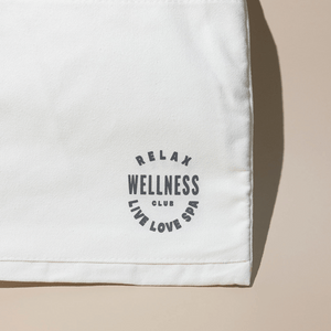 Close-up of a cream-colored fabric tote bag corner with a circular black logo reading "Relax Wellness Club Live Love Spa," part of the Wellness Club Mini Tote | Live Love Spa collection.
