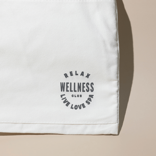 Close-up of a cream-colored fabric tote bag corner with a circular black logo reading "Relax Wellness Club Live Love Spa," part of the Wellness Club Mini Tote | Live Love Spa collection.