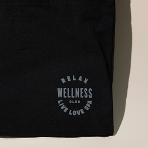 Close-up of the black fabric corner of the Wellness Club Mini Tote | Live Love Spa featuring light gray text that reads "RELAX WELLNESS CLUB LIVE LOVE SPA" on a neutral background.