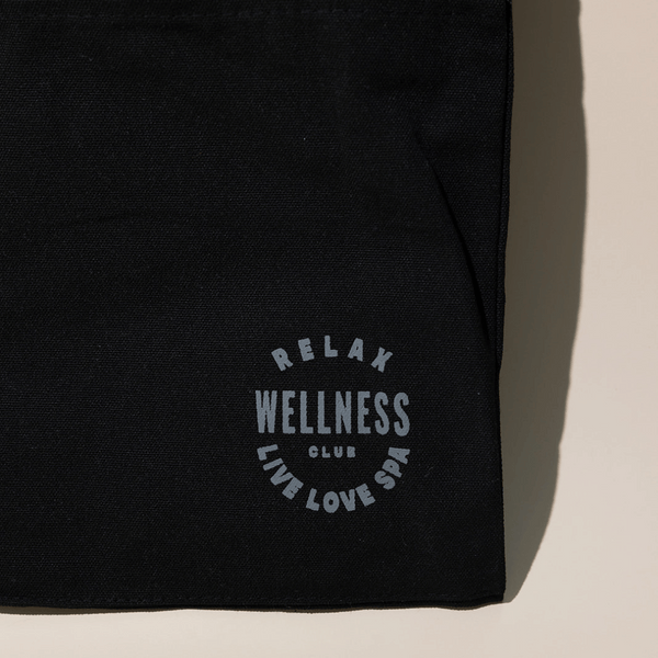 Close-up of the black fabric corner of the Wellness Club Mini Tote | Live Love Spa featuring light gray text that reads "RELAX WELLNESS CLUB LIVE LOVE SPA" on a neutral background.