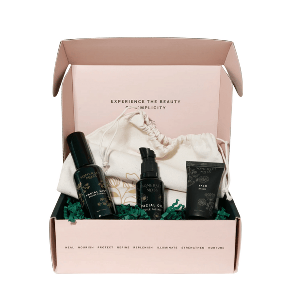Open gift box containing a skincare set with facial mist, facial oil, and balm tubes arranged on green shredded paper and a white drawstring pouch, showcasing the Core Collection Kit | Somerset Moss.