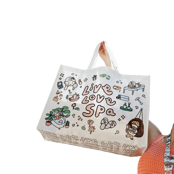 A white tote bag featuring cute spa-themed illustrations and the text "Live Love Spa," held by a person wearing an orange knit top, showcasing the Live Love Spa Kawaii Event Bag | Live Love Spa.