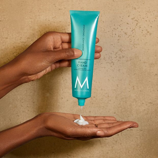 A person dispensing white cream from a teal tube labeled Hand Cream | Moroccanoil onto their palm, shown against a neutral textured background.