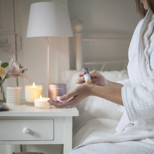 Person in a white bathrobe applying Sleep Well Therapy Balm | Scentered on their wrist beside lit candles on a bedside table creating a calming wellness and skincare routine atmosphere.