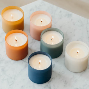 Six lit candles in frosted glass holders in soft pastel colors arranged on a marble surface, featuring Aspen + Fog Dignity Series Glass Jar Soy Candle | Calyan Wax Co. - 5.3 oz.