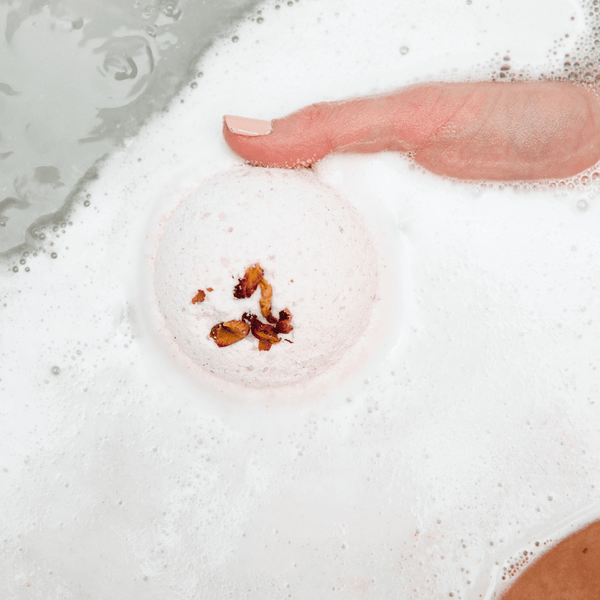 Hand gently holding a foamy, white bath bomb with dried flower petals on its surface, partially submerged in a bath, showcasing Mama's Perch Bath Bomb | Bathorium for a soothing skincare and wellness experience.