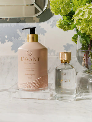 Elegant bathroom scene with a blush-colored hand soap and clear fragrance bottle displayed on a marble countertop next to a vase of green hydrangeas, featuring Room Spray | L'AVANT Collective.