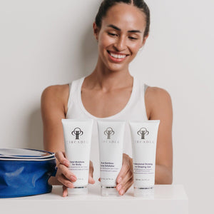 A smiling woman wearing a white tank top displays three skincare tubes from the Body Regimen Bundle | Circadia next to a blue cosmetic bag on a white surface.