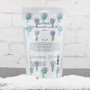 Packaging of Lavender Bubbling Bath Salts | Primal Elements featuring lavender illustrations, set against a white brick background with bath salts scattered in front.