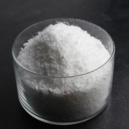 Close-up of white granular Sore Muscle Bubbling Bath Salts | Primal Elements in a clear glass container for body care and wellness use.