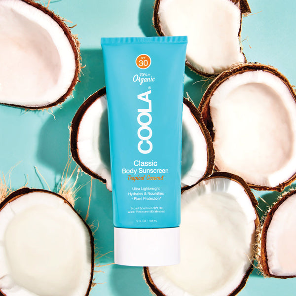 Classic Body Organic Sunscreen Lotion SPF 30 - Tropical Coconut | COOLA tube surrounded by coconut halves on a light blue background, highlighting natural skincare and body care elements.