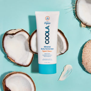 Tube of Mineral Body Organic Sunscreen Lotion SPF 30 - Tropical Coconut | COOLA on a blue surface surrounded by halved coconuts and sunscreen lotion swatch, highlighting natural skincare and body care.