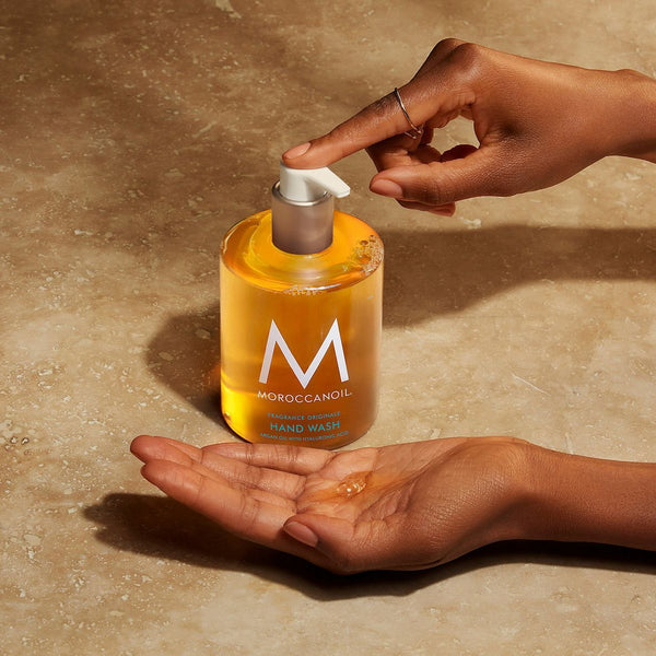 A person dispensing golden liquid from a bottle of Hand Wash | Moroccanoil into their palm on a beige textured surface, highlighting skincare and body care.
