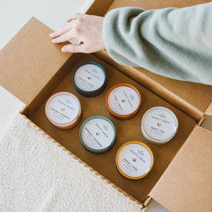 Person opening a cardboard box containing six colorful candles from the Evergreen + Eucalyptus Dignity Series Glass Jar Soy Candle | Calyan Wax Co. - 5.3 oz collection.