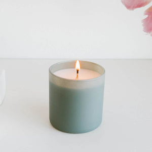 Lit candle in a soft green glass jar on a white surface with a minimal background, showcasing the Evergreen + Eucalyptus Dignity Series Glass Jar Soy Candle | Calyan Wax Co. - 5.3 oz.