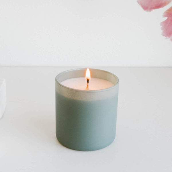 Lit candle in a soft green glass jar on a white surface with a minimal background, showcasing the Evergreen + Eucalyptus Dignity Series Glass Jar Soy Candle | Calyan Wax Co. - 5.3 oz.