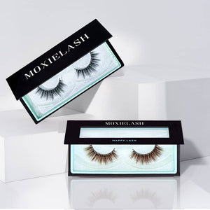 Two pairs of false eyelashes in elegant black and teal packaging labeled Happy Lash | Moxielash, displayed on white geometric blocks in a clean, minimal setting.