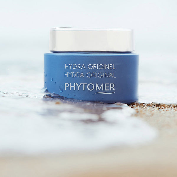 Hydra Original Cream | Phytomer in a blue jar with a white lid sitting on a sandy beach with gentle waves, highlighting its skincare and hydration benefits.