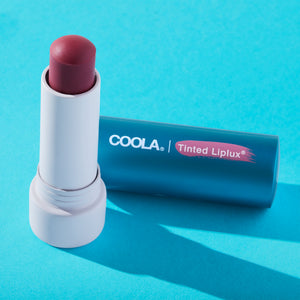 Open tube of Classic Liplux® Lip Balm Tinted SPF 30 | COOLA with muted pink tint resting on a blue surface, highlighting skincare lip protection and wellness.