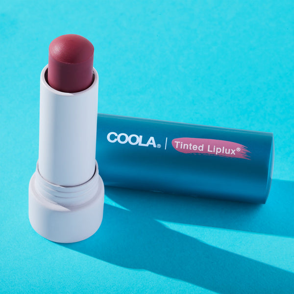 Open tube of Classic Liplux® Lip Balm Tinted SPF 30 | COOLA with muted pink tint resting on a blue surface, highlighting skincare lip protection and wellness.
