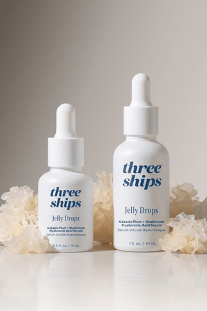 Two white dropper bottles of Jelly Drops™ Kakadu Plum + Mushroom Hyaluronic Acid Serum | Three Ships surrounded by translucent natural mushroom-like elements on a neutral background, showcasing skincare serum packaging in 15 ml and 30 ml sizes.