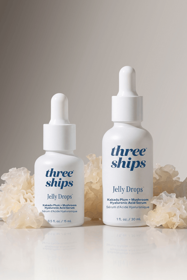 Two white dropper bottles of Jelly Drops™ Kakadu Plum + Mushroom Hyaluronic Acid Serum | Three Ships surrounded by translucent natural mushroom-like elements on a neutral background, showcasing skincare serum packaging in 15 ml and 30 ml sizes.
