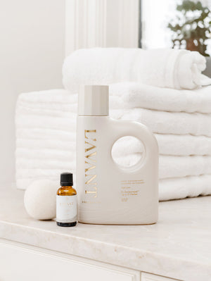 Bottle of High Performing Laundry Detergent - Fresh Linen | L'AVANT Collective placed on a bathroom counter with neatly stacked white towels and a small essential oil bottle beside it in a clean, bright setting.