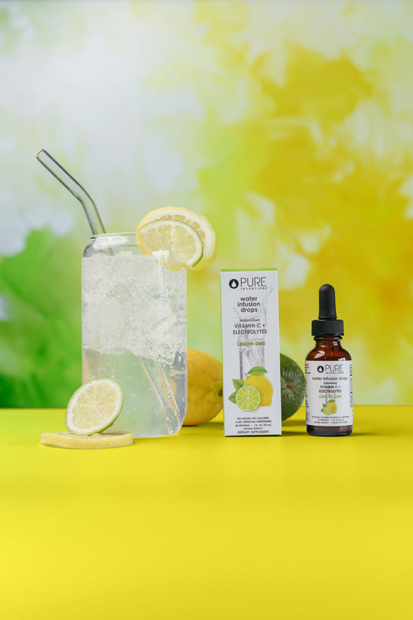 Bottle and box of Vitamin C + Electrolytes Lemon-Lime Water Infusion Drops - 30 servings | Pure Inventions next to a glass of sparkling water with lemon slices and a metal straw on a yellow table.