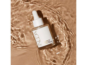 Clear glass dropper bottle of Hyaluronic Self Tanning Serum | NUDA resting on shimmering, warm-toned water surface for skincare and body care wellness.