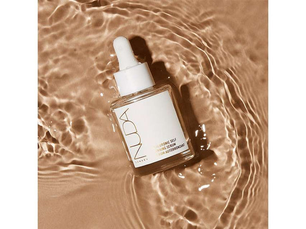 Clear glass dropper bottle of Hyaluronic Self Tanning Serum | NUDA resting on shimmering, warm-toned water surface for skincare and body care wellness.