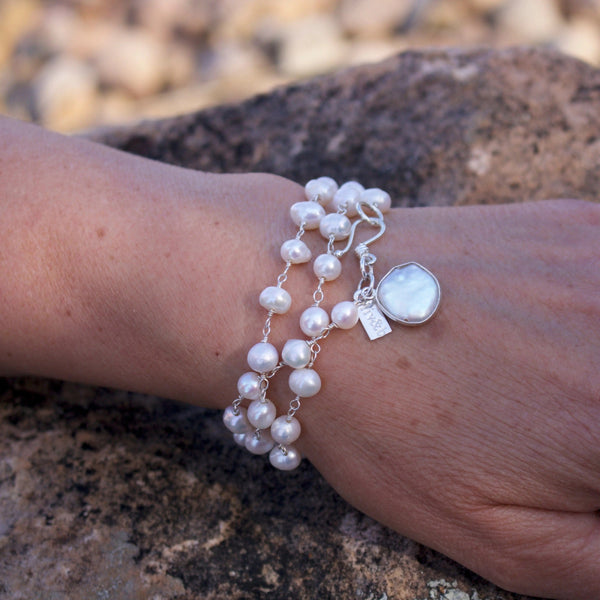 It's A Wrap Triple Pearl Bracelet | Liv & B featuring three strands of white pearls wrapped around the wrist with a silver charm against a natural stone background.