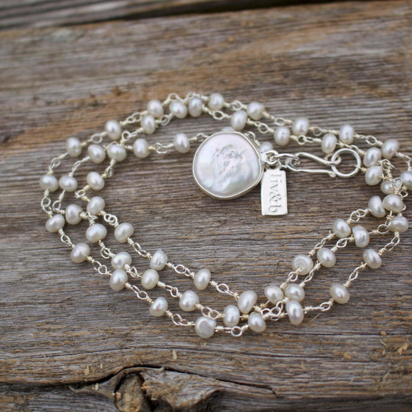 It's A Wrap Triple Pearl Bracelet | Liv & B featuring delicate white pearls on silver chains with a central coin-shaped pearl charm and a silver clasp on a wooden surface.