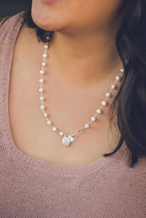 Close-up of a person wearing a delicate pearl necklace featuring a clasp with a charm, styled elegantly with a soft pink knit top, showcasing the It's A Wrap Triple Pearl Bracelet | Liv & B.
