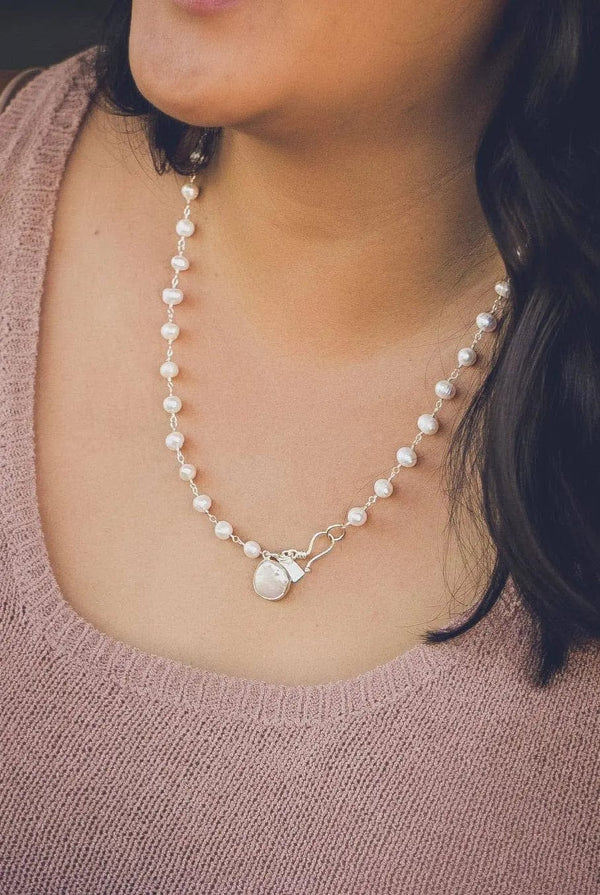 Close-up of a person wearing a delicate pearl necklace featuring a clasp with a charm, styled elegantly with a soft pink knit top, showcasing the It's A Wrap Triple Pearl Bracelet | Liv & B.