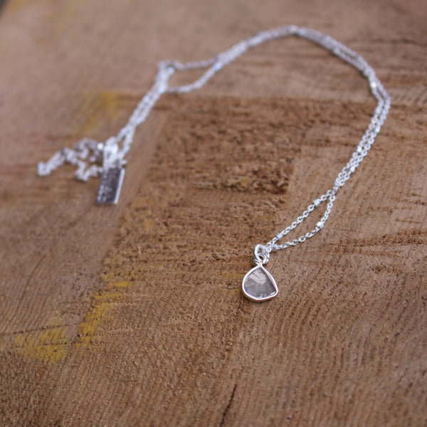 Diamond Slice Sterling Silver Necklace | Liv & B featuring a delicate silver chain with a small, uniquely shaped diamond slice pendant resting on a textured wooden surface.