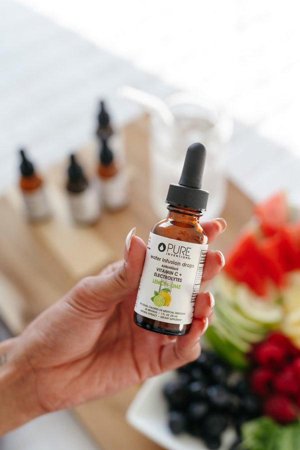Hand holding a bottle of Vitamin C + Electrolytes Lemon-Lime Water Infusion Drops - 30 servings | Pure Inventions with a blurred background of fruits and additional bottles on a wooden tray.