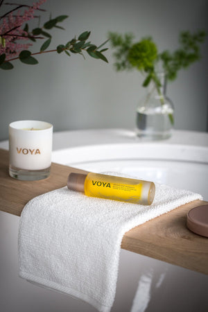 Moonlight Moments Bath & Shower Oil | VOYA displayed on a white towel over a wooden bath ledge, accompanied by a VOYA candle and a glass vase with green foliage, creating a serene spa atmosphere.
