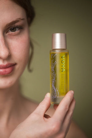 A close-up of a person holding a clear bottle filled with golden oil labeled Moonlight Moments Bath & Shower Oil | VOYA, showcasing skincare and body care product for wellness.