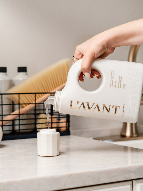 Hand pouring clear liquid from a white bottle labeled L'AVANT into a white measuring cup on a kitchen countertop, showcasing High Performing Laundry Detergent - Fresh Linen | L'AVANT Collective.