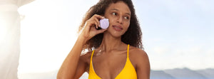 Woman in yellow swimsuit using a purple BEAR™ 2 Go | FOREO facial massage device outdoors with a clear sky background, promoting skincare and wellness.