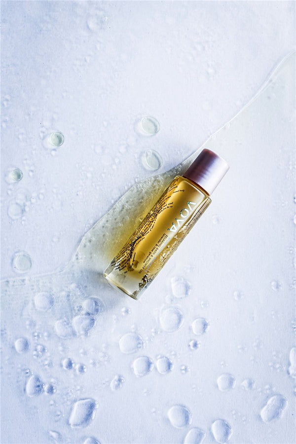 Moonlight Moments Bath & Shower Oil | VOYA bottle lying on a wet surface with clear water droplets, emphasizing skincare and body care essentials for a relaxing wellness routine.