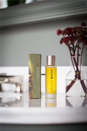 Moonlight Moments Bath & Shower Oil | VOYA in a yellow bottle with a rose gold cap next to its green packaging, displayed on a bathroom countertop with red flowers in a glass vase.