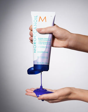 Hands dispensing vibrant purple conditioner from a tube of Blonde Perfecting Purple Conditioner | Moroccanoil against a neutral background.