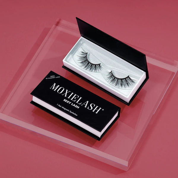 Open black box containing a pair of dramatic, voluminous false eyelashes labeled Sexy Lash | Moxielash, displayed on a pink surface.