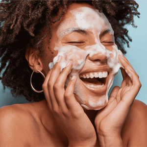 Smiling woman with natural curly hair gently cleansing her face with rich foam, illustrating the fresh and soothing effect of Gentle Foaming Cleanser | Privai for skincare.