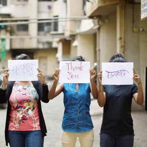 Three people standing outdoors holding signs in front of their faces with handwritten messages, featuring the "Brass Droplet Earrings | Purpose Jewelry" on their ears.